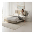 Person sitting on a beige upholstered bed in a bright bedroom