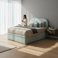 Person seated on mint upholstered bed beside a window